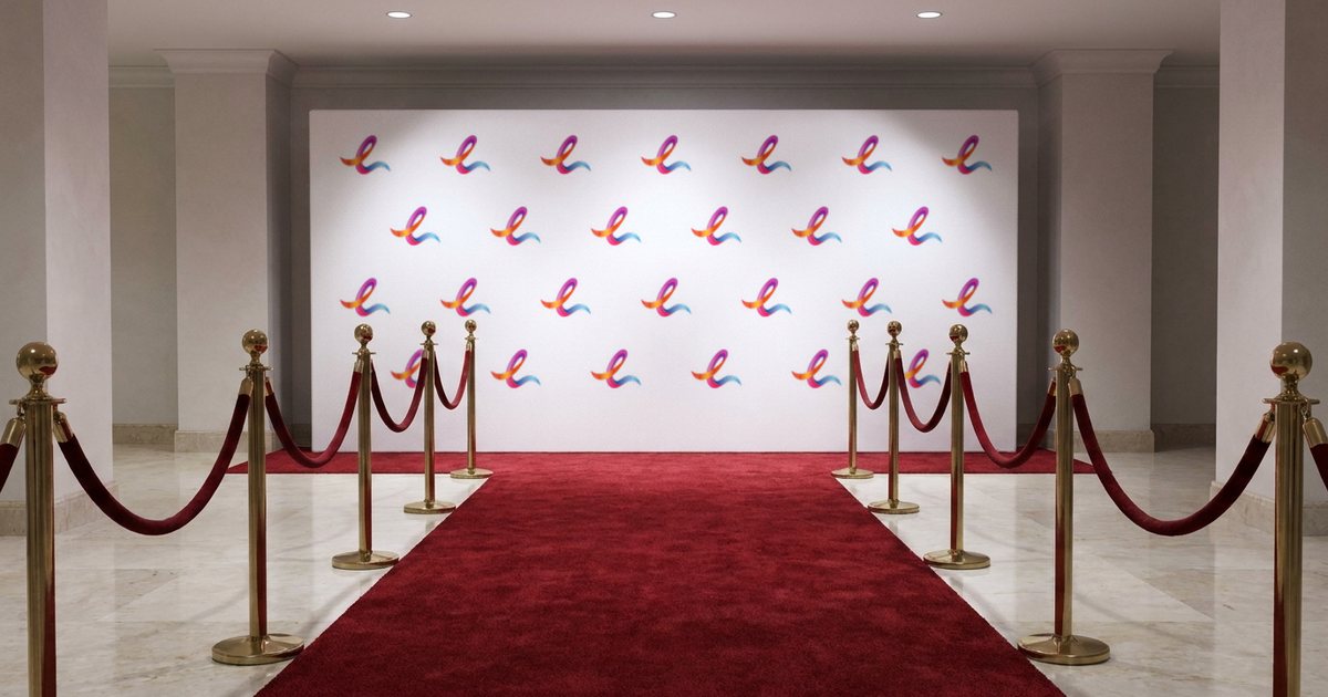 Step-and-repeat media wall printed with a grid of EZ Print ribbon marks behind a red carpet and brass stanchions.
