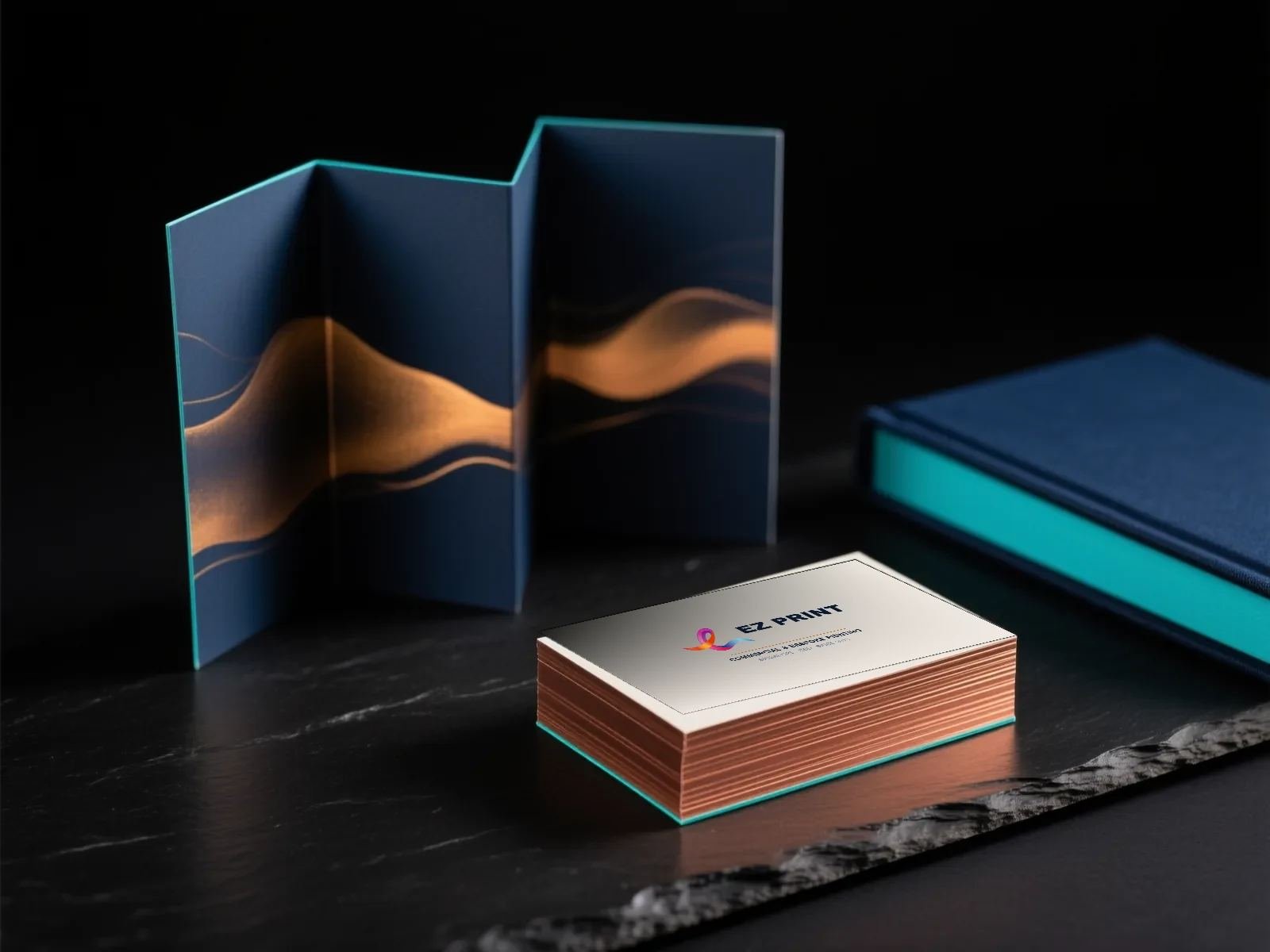 A stack of cream business cards printed with the EZ Print ribbon mark and edged in copper, beside a concertina-folded navy brochure and a navy cloth-bound book.